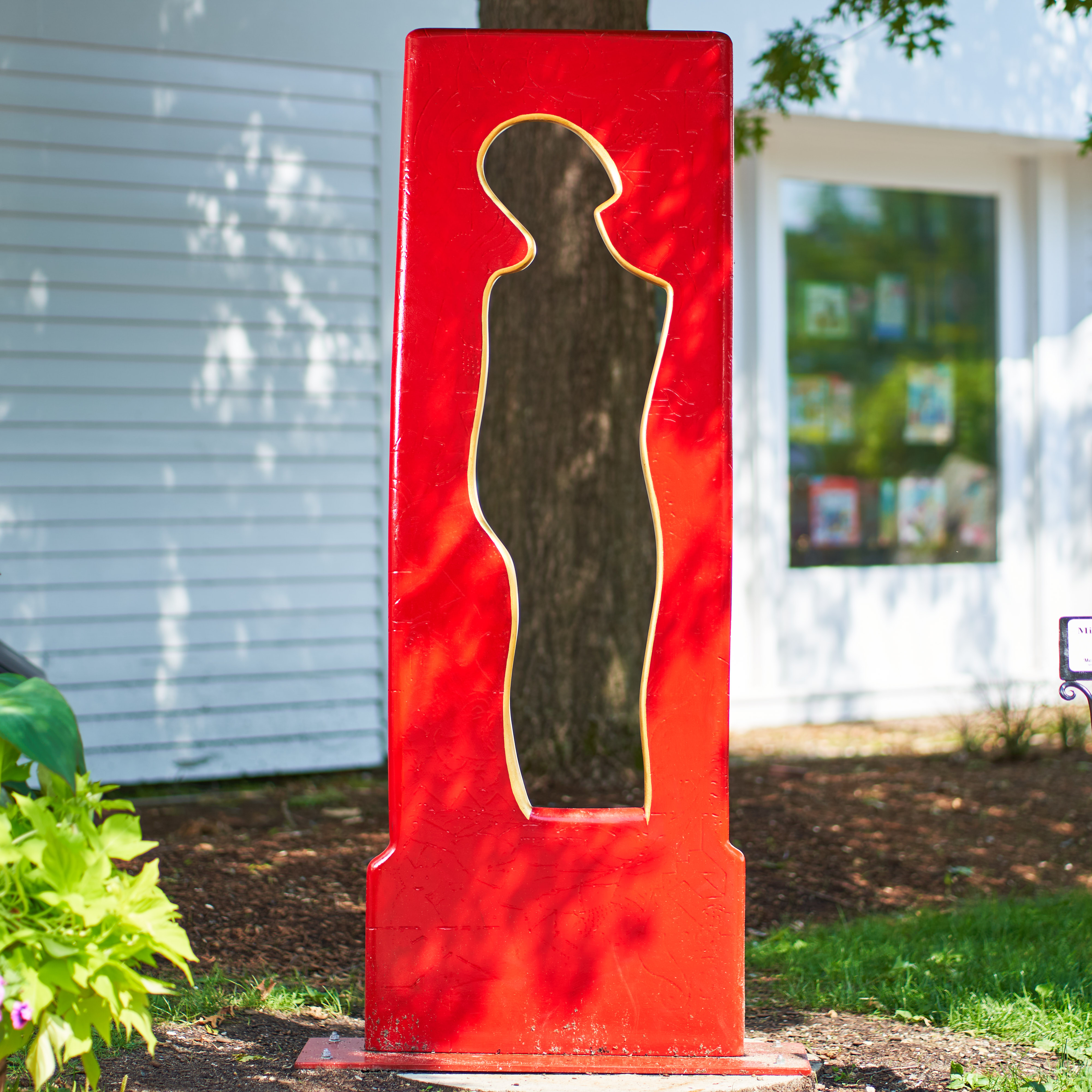 Sculpture of the negative space around an abstract human form. It is bright red with gold along the edge of the center void. Painted urethane and gold leaf.