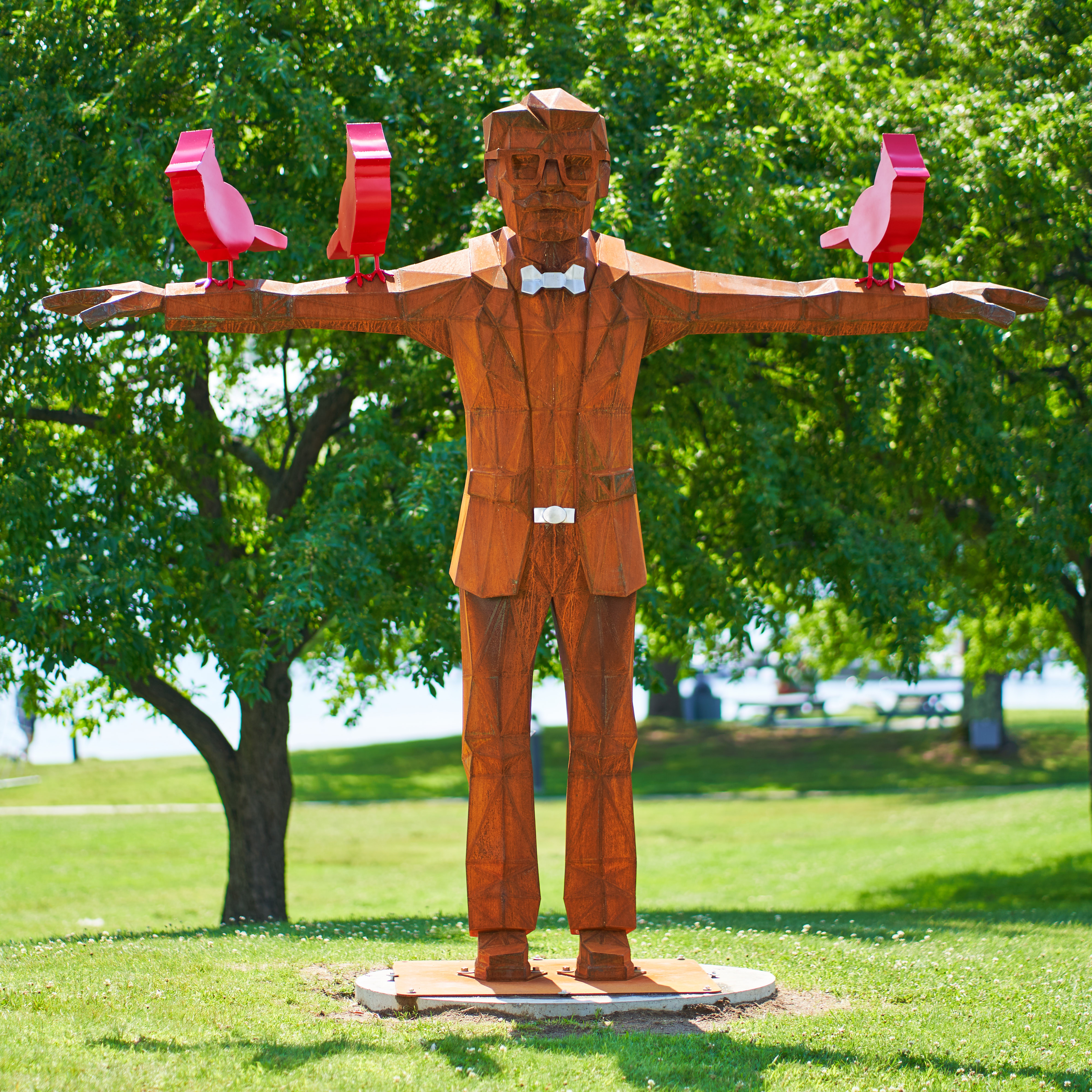Sculpture of a man facing the camera with arms outstretched. Three red birds rest on his arms.